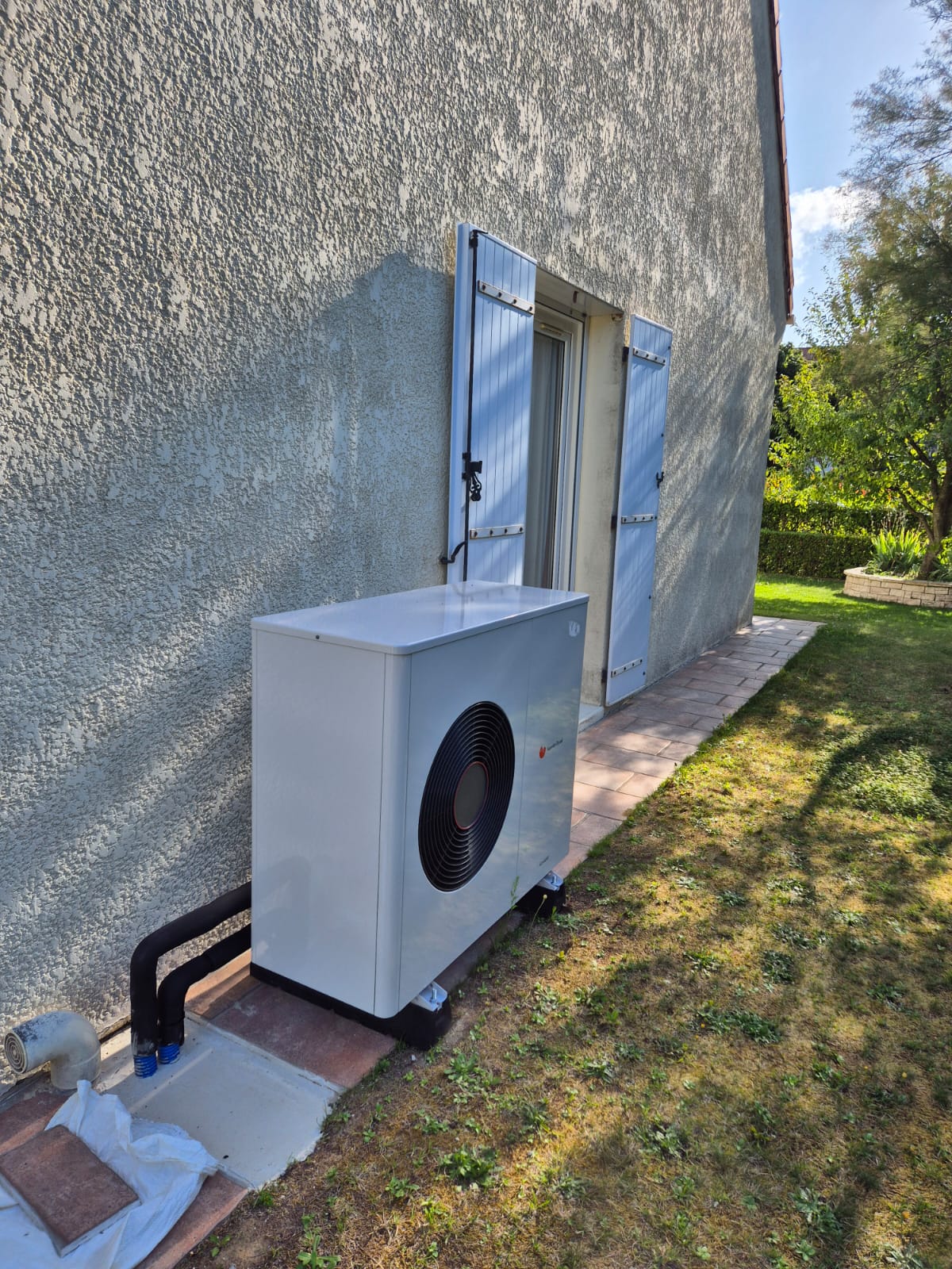 Pompe à chaleur Saunier Duval blanche posée sur plots le long d'une façade crépie, près d'une porte-fenêtre