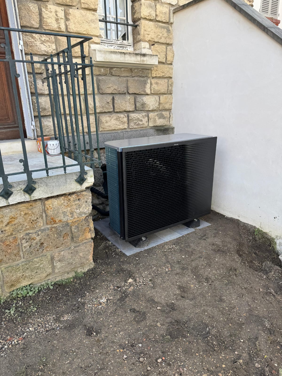 Pompe à chaleur Daikin haute température posée sur dalle au pied d'une maison en pierre, à côté du perron